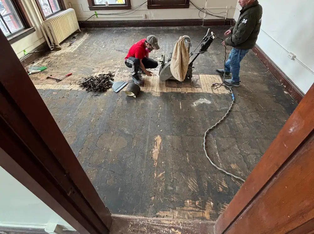 Two Dream Home crew members pulling up an old floor in a room with tall windows