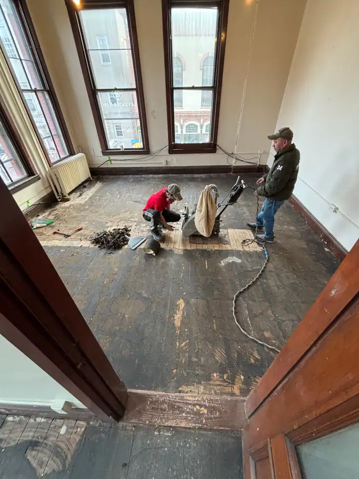 Two crew members pulling up an old floor in a room with tall windows